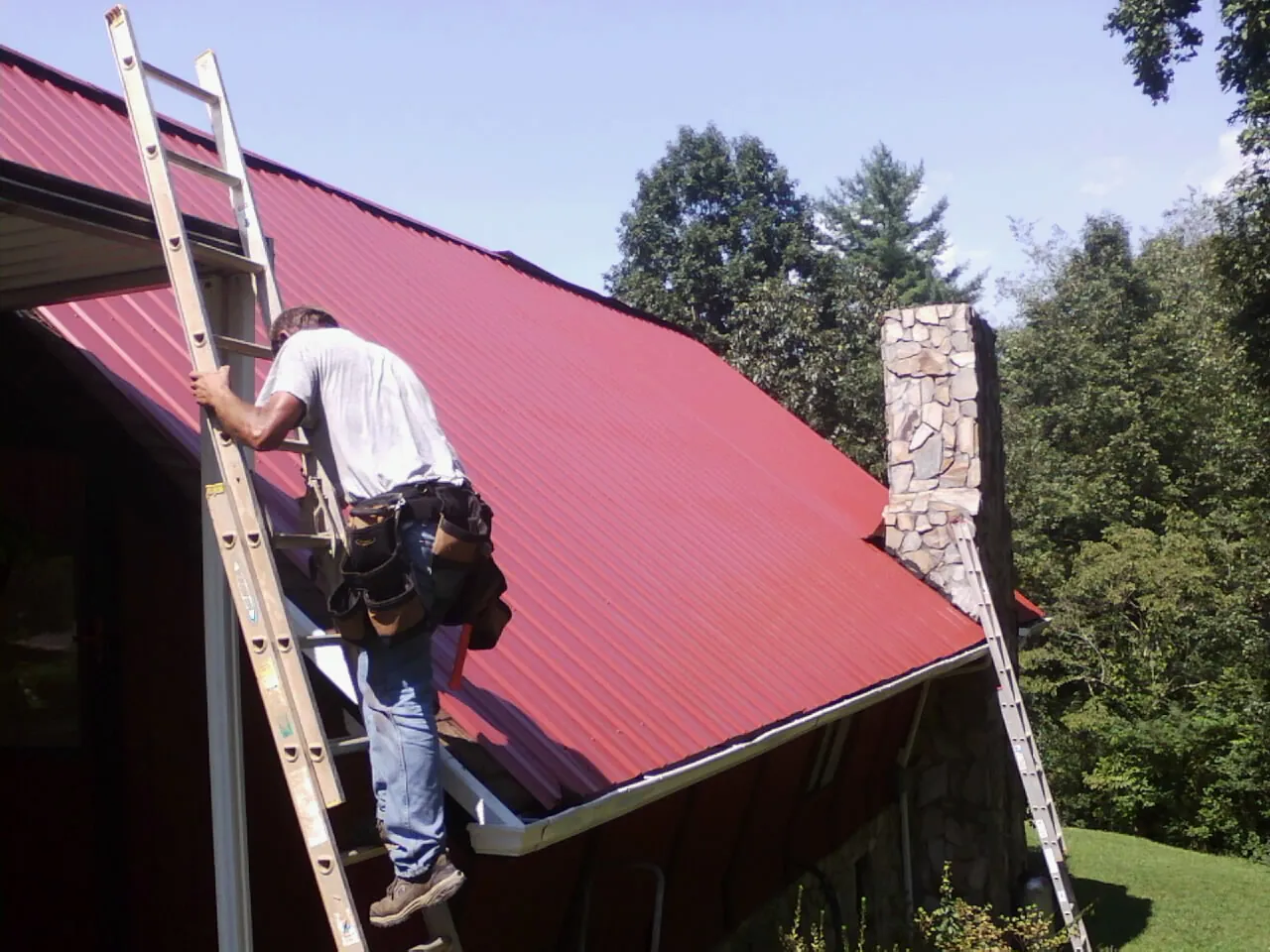 Roofer on a ladder beside a red metal roof and stone chimney — Soffit & Fascia Repair by Roofing Brothers in East Galesburg